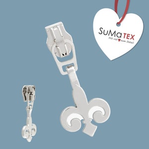 Könnte beinhalten: Silberner Reißverschluss-Zipper mit einem Fleur-de-lis-Design. Der Zipper ist an einem Metall-Schieber mit einer kleinen Schlaufe befestigt. Der Markenname "SUMATEX" ist auf einem weißen herzförmigen Etikett mit dem Text "Mit viel zum Detail" gedruckt.