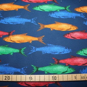 Op de afbeelding: Een blauwe stof met een herhalend patroon van kleurrijke vissen. De vissen zijn in tinten rood, oranje, groen en blauw. Een houten liniaal met markeringen in centimeters ligt op de stof.