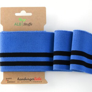 May include: Blue fabric with two black stripes, displayed with a brown cardboard tag. The tag has the text "ALB Stoffe" and "hamburgerliebe by Susanne Firmenich" along with "MADE IN GERMANY". The fabric is folded.