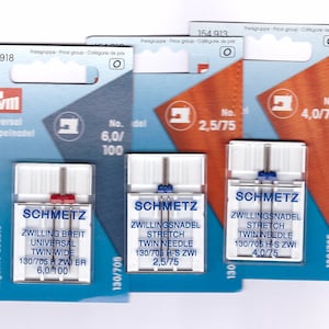 May include: Three packets of sewing machine needles. The packets are blue, orange, and white, with the brand name SCHMETZ. The packets have text in German and English, including "Twin Needle" and needle sizes.