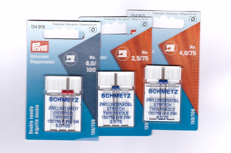 May include: Three packets of sewing machine needles. The packets are blue, orange, and white, with the brand name SCHMETZ. The packets have text in German and English, including "Twin Needle" and needle sizes.