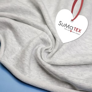 May include: Close-up of light grey fabric with visible texture and folds. A white heart-shaped tag with the text "SuMaTEX" and "Mit viel zum Detail" is attached with a red ribbon. The fabric is draped over a blue surface.