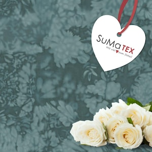 Op de afbeelding: Een wit hartvormig label met de tekst "SUMATEX" en "Mit viel zum Detail" in zwarte letters. Het label hangt aan een rood lint en is geplaatst op een blauwe en witte bloemenachtergrond. Een boeket witte rozen bevindt zich in de rechterbenedenhoek van de afbeelding.