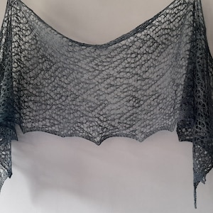 Può includere: Un scialle di uncinetto grigio con un motivo a pizzo. Lo scialle è drappeggiato su una parete bianca.