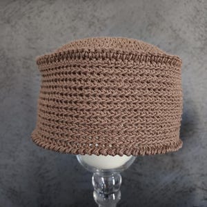 May include: A brown crocheted hat with a textured pattern. The hat is sitting on a clear glass pedestal.