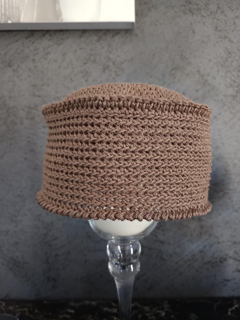 May include: A brown crocheted hat with a textured pattern. The hat is sitting on a clear glass pedestal.