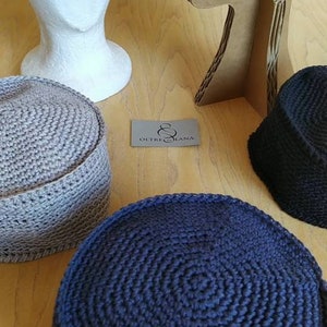 May include: Four crocheted hats in different colours and styles. The hats are displayed on mannequins and a wooden surface. The hats are made of a soft, textured yarn. The label "Oltre Lana" is visible in the image.