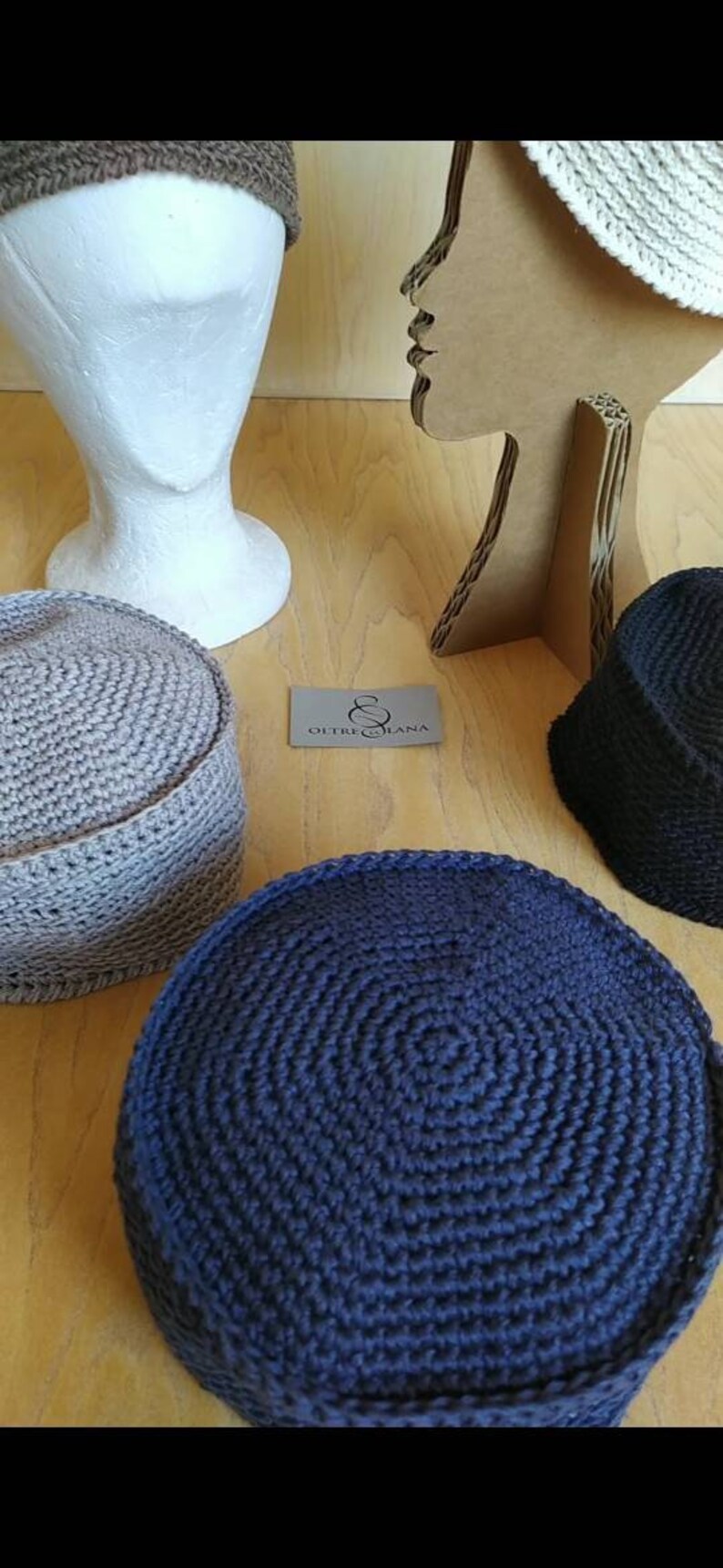 May include: Four crocheted hats in different colours and styles. The hats are displayed on mannequins and a wooden surface. The hats are made of a soft, textured yarn. The label "Oltre Lana" is visible in the image.