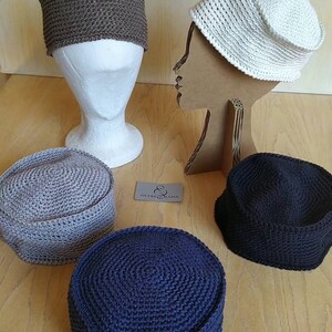 May include: Four crocheted hats in different colours: brown, white, grey, and dark blue. The hats are displayed on mannequins and a wooden surface. The text "OUTRE LANA" is visible on a tag.
