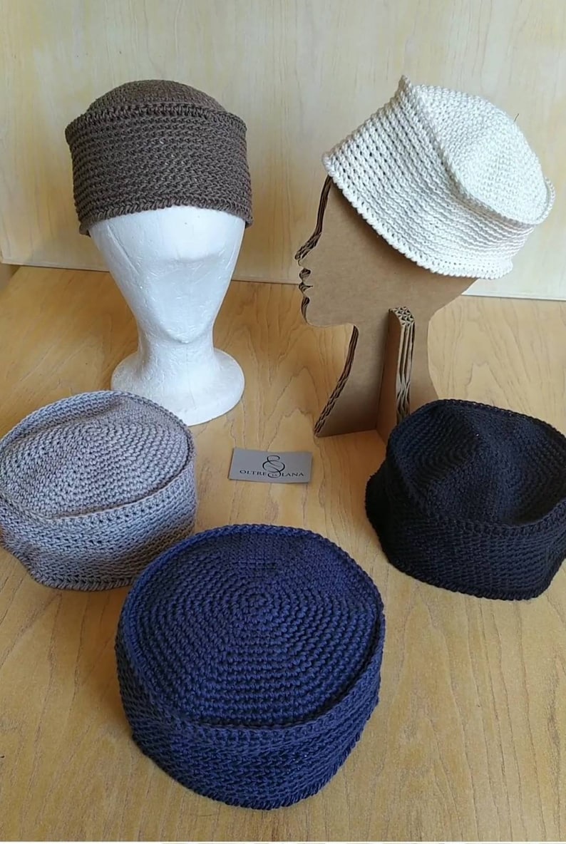 May include: Four crocheted hats in different colours: brown, white, grey, and dark blue. The hats are displayed on mannequins and a wooden surface. The text "OUTRE LANA" is visible on a tag.