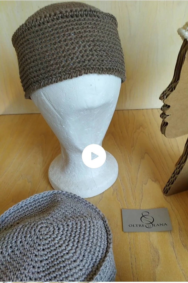 May include: Two brown crocheted hats on a mannequin head and a wooden surface. The hats are made with a thick yarn and have a simple design. The text "OLTRE LA LANA" is visible on a small card.