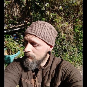 May include: A brown knitted beanie with a textured pattern. The beanie is worn by a person with a beard and a brown jacket.