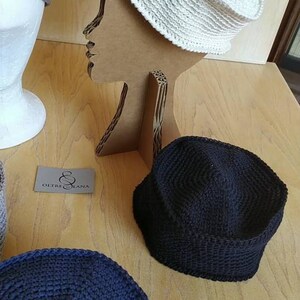 May include: Three crocheted hats in different colours: beige, black and navy blue. The hats are displayed on a wooden surface and a cardboard mannequin.