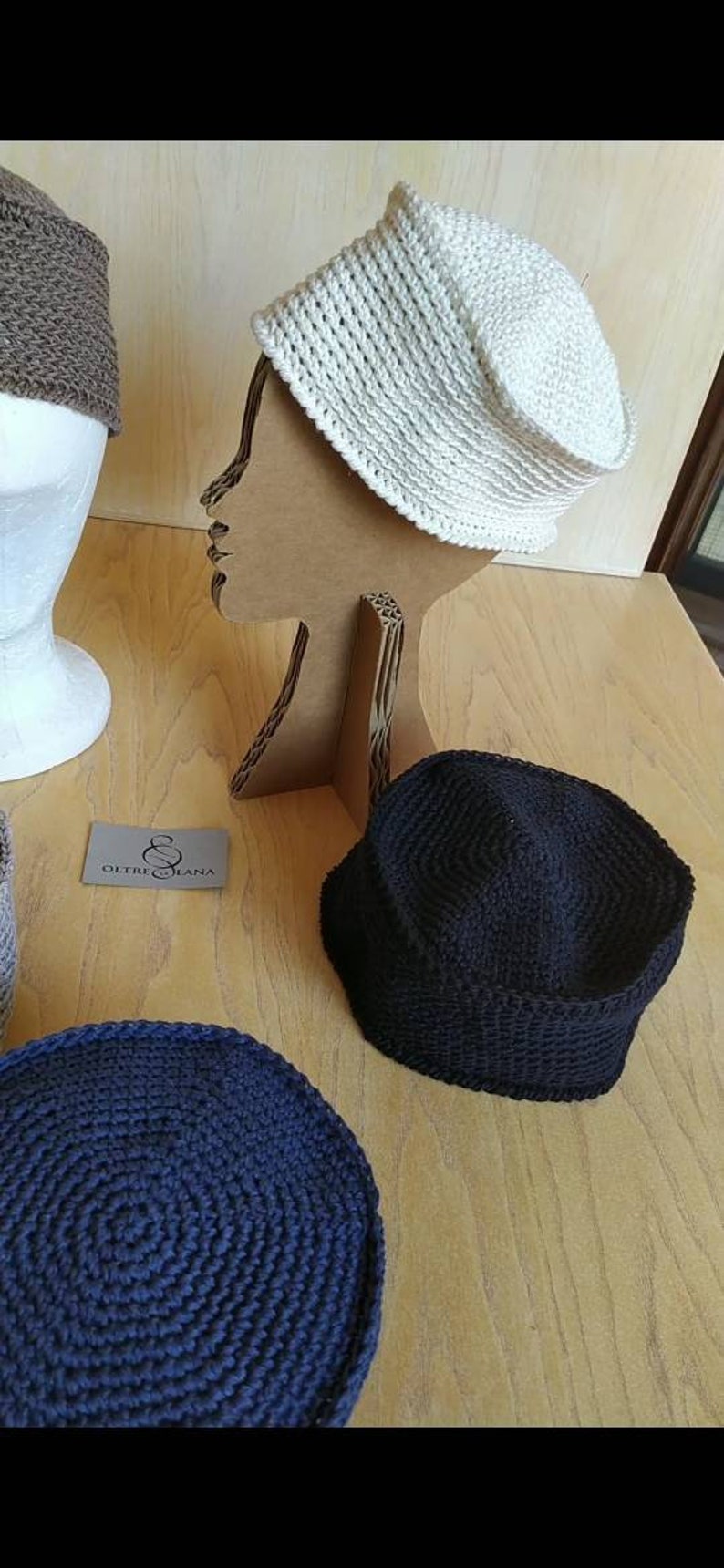 May include: Three crocheted hats in different colours: beige, black and navy blue. The hats are displayed on a wooden surface and a cardboard mannequin.