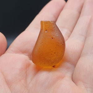 May include: A small, amber-coloured glass vase with a rounded, teardrop shape. The vase has a textured surface and a small opening at the top.