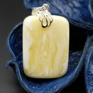Könnte beinhalten: Ein rechteckiger Bernstein-Anhänger mit einer silbernen Öse. Der Bernstein ist hellgelb mit weißen Streifen. Der Anhänger liegt auf einem blauen Hintergrund.