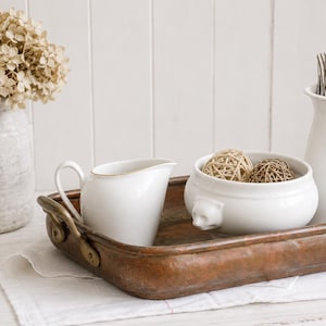 Op de afbeelding: Een rechthoekig, gehamerd koperen dienblad met messing handgrepen bevat een witte keramische melkkan, een witte keramische kom met leeuwenkopaccenten en twee gevlochten bollen, en een witte keramische kan met zilveren bestek.