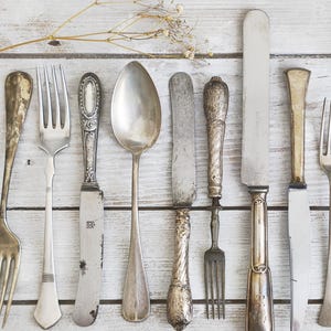 Könnte beinhalten: Eine Auswahl an antikem Besteck, darunter Gabeln, Messer und Löffel, angeordnet auf einer verwitterten, hellen Holzoberfläche. Das Besteck hat verzierte Griffe und eine Vintage-Ästhetik, perfekt für eine rustikale Tischdekoration.
