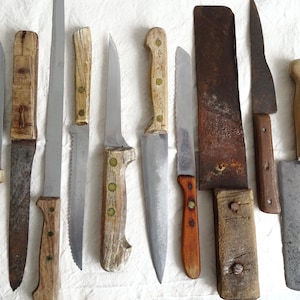 Könnte beinhalten: Eine Sammlung verschiedener Vintage-Messer mit Holz- und Metallgriffen, darunter ein großes Hackmesser. Die Klingen weisen Gebrauchsspuren auf, was darauf hindeutet, dass sie benutzt wurden. Die Messer sind auf einem weißen Tuch angeordnet.