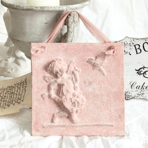 Peut inclure: Un tableau mural carré rose avec un bas-relief d'un chérubin et d'un oiseau. Le chérubin est représenté avec des ailes et un arc. La suspension a une poignée en ruban rose. Une statue de chérubin blanc et un bougeoir sont en arrière-plan.