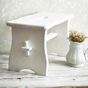 Puede incluir: Un taburete de madera blanco con un asiento rectangular y un detalle en forma de cruz en el lateral. El taburete está pintado de blanco y tiene una estética rústica. Una pequeña jarra blanca con flores secas está cerca.