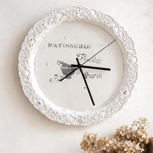 Könnte beinhalten: Eine runde Wanduhr mit einem weißen, verzierten und geprägten Rahmen mit floralen Details. Das Zifferblatt ist weiß mit schwarzen Zeigern und schwarzer Schrift, die Patisserie, Chocolate und Paris liest, sowie einer Grafik von Mörser und Stößel.