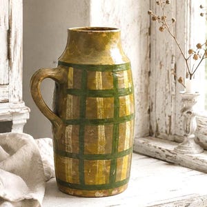 Puede incluir: Una jarra cilíndrica de cerámica con un patrón de cuadrícula en color amarillo mostaza y verde oscuro. El recipiente tiene una sola asa curva en el lateral, un cuello ancho y un acabado esmaltado liso con texturas de pinceladas visibles.