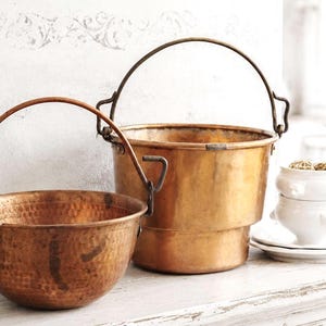 May include: Two copper vessels with metal handles. The smaller vessel has a hammered texture and a rounded base. The larger vessel is taller with a smooth finish and a stepped base. Both items feature arched handles attached to the sides.