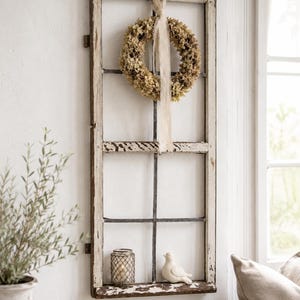 Puede incluir: Un marco de ventana rectangular alto de madera blanca desgastada montado en una pared. Una corona de hortensias secas con una cinta de tela larga y clara cuelga de la parte superior. Un pequeño farol de metal y un pájaro de cerámica blanca descansan en el borde inferior.