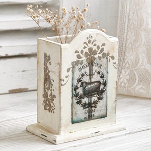 May include: A rectangular wooden container with a distressed white finish, featuring a central graphic of a sheep surrounded by a wreath. The text reads SHEEP WOOL, Natural, and FARM est. 1845. Dried baby's breath flowers are placed inside the container.