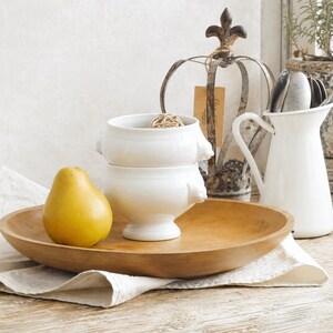 May include: A large, round, light brown wooden bowl containing a single yellow pear and two stacked white ceramic bowls with small side handles. The bowls are positioned near a white metal pitcher holding silver spoons and a metal crown ornament.
