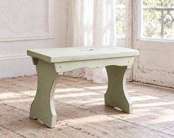 Peut inclure: Un petit banc rectangulaire en bois peint en vert sauge pâle avec une texture vieillie et usée. Le banc présente une surface supérieure plate et deux larges supports latéraux incurvés reliés par une traverse horizontale sous le siège.
