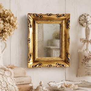 Puede incluir: Un espejo de pared rectangular con un marco dorado envejecido y ornamentado con detalles de volutas intrincadas. El cristal del espejo tiene un aspecto ligeramente envejecido y moteado, reflejando el espacio interior circundante.
