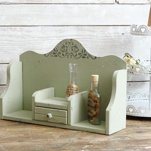May include: A distressed, sage green wooden shelf with a small drawer and decorative top. Two glass bottles with cork stoppers sit on the shelf. A white tiered stand is in the background.
