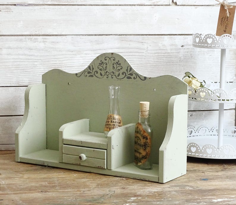 May include: A distressed, sage green wooden shelf with a small drawer and decorative top. Two glass bottles with cork stoppers sit on the shelf. A white tiered stand is in the background.