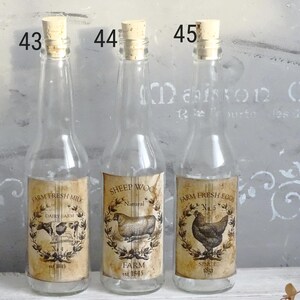 Puede incluir: Tres botellas de vidrio transparente con tapones de corcho, etiquetadas 43, 44 y 45. Cada botella presenta una etiqueta de papel de estilo vintage: Farm Fresh Milk con una vaca, Sheep Wool con una oveja y Farm Fresh Eggs con una gallina, todo en tonos sepia.