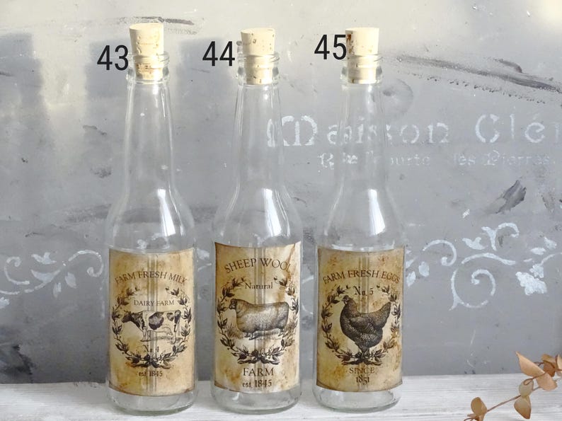 Puede incluir: Tres botellas de vidrio transparente con tapones de corcho, etiquetadas 43, 44 y 45. Cada botella presenta una etiqueta de papel de estilo vintage: Farm Fresh Milk con una vaca, Sheep Wool con una oveja y Farm Fresh Eggs con una gallina, todo en tonos sepia.