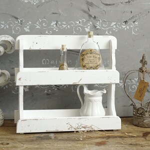 May include: A two-tiered white wooden shelf with a distressed finish. On the top shelf sit two glass bottles with cork stoppers. The bottom shelf holds a small white ceramic pitcher. The shelf is displayed alongside a white fleur-de-lis and a metal crown.