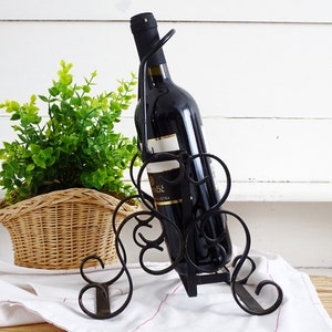 Pode incluir: Um porta-garrafas de vinho de metal preto com uma garrafa de vinho tinto dentro. O porta-garrafas tem um design decorativo em espiral. O porta-garrafas está sentado sobre um pano branco.