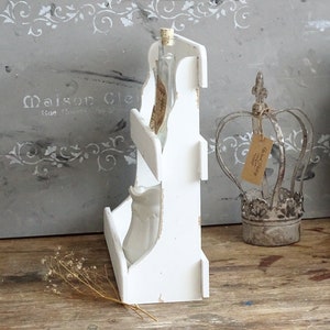 May include: A white wooden tiered shelf with a vertical profile, featuring two open compartments. The top shelf holds a clear glass bottle with a cork stopper and a paper label, while the bottom shelf contains a small white ceramic pitcher.