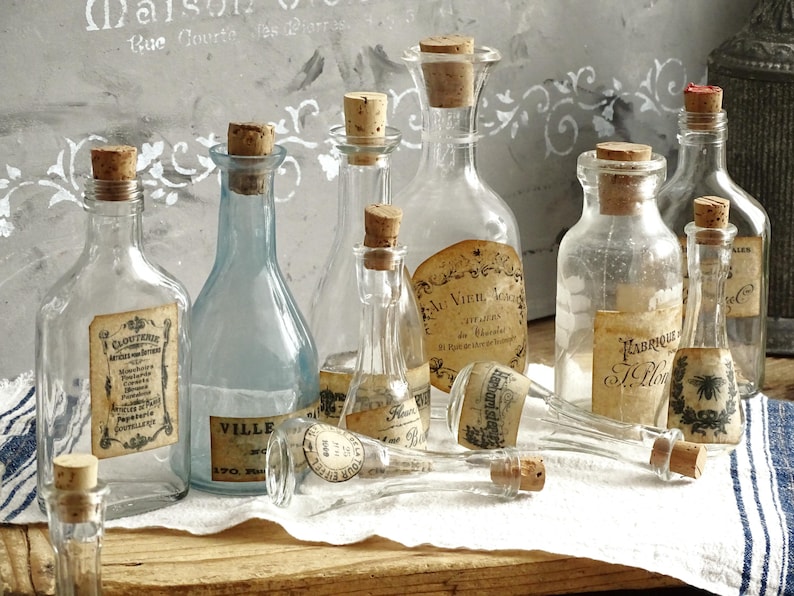 Puede incluir: Una colecci&oacute;n de botellas de vidrio transparente y azul claro de diversas formas y tama&ntilde;os, cada una con un tap&oacute;n de corcho. Varias botellas presentan etiquetas de papel envejecido con texto impreso y gr&aacute;ficos sobre una tela blanca.