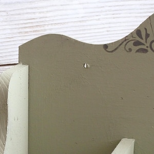 May include: Close-up of a wooden shelf with a decorative design. The shelf is painted in a muted green color, with a white accent. The top edge features a stenciled floral pattern in a darker shade of green.