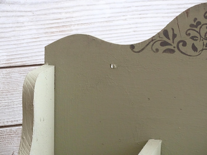 May include: Close-up of a wooden shelf with a decorative design. The shelf is painted in a muted green color, with a white accent. The top edge features a stenciled floral pattern in a darker shade of green.