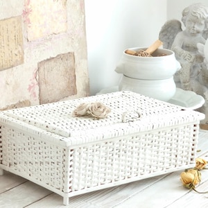 May include: A white rectangular woven box with a hinged lid, adorned with a fabric flower. The box is on a white wooden surface, with a decorative background featuring vintage paper and floral patterns. Dried roses are nearby.