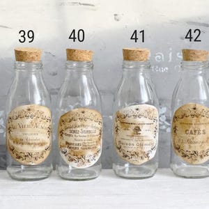 Puede incluir: Cuatro botellas de vidrio transparente con tapones de corcho, cada una con una etiqueta de papel de estilo antiguo. Las etiquetas est&aacute;n numeradas 39, 40, 41 y 42, mostrando varios textos en franc&eacute;s como Au Vieil Acacia y Caf&eacute;s Launay.