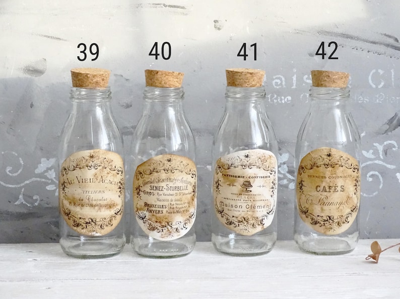 Puede incluir: Cuatro botellas de vidrio transparente con tapones de corcho, cada una con una etiqueta de papel de estilo antiguo. Las etiquetas est&aacute;n numeradas 39, 40, 41 y 42, mostrando varios textos en franc&eacute;s como Au Vieil Acacia y Caf&eacute;s Launay.