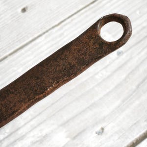 Puede incluir: Un mango de metal largo, estrecho y plano con una textura de superficie marr&oacute;n oscura, muy oxidada y con &oacute;xido. El extremo del mango presenta un orificio circular, dise&ntilde;ado para colgar o almacenar, que muestra signos significativos de desgaste.