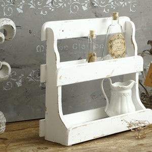 May include: A two-tiered white wooden shelf with a distressed finish. The top shelf holds two clear glass bottles with cork stoppers and labels. The bottom shelf holds a white ceramic pitcher with a handle and a decorative scalloped rim detail.