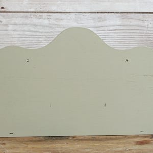 May include: A sage green wooden wall shelf with a scalloped top edge. The shelf is rectangular with a flat back and is mounted on a wooden surface. The background is a light-colored wooden wall.