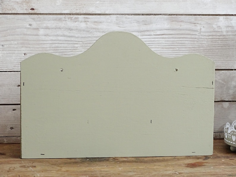 May include: A sage green wooden wall shelf with a scalloped top edge. The shelf is rectangular with a flat back and is mounted on a wooden surface. The background is a light-colored wooden wall.